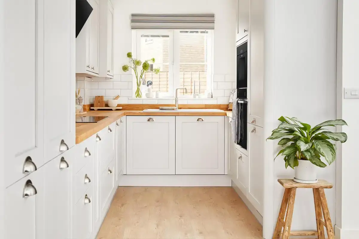 آشپزخانه با کابینت‌ها و دیوار سفید و پارکت‌های رنگ چوب به همراه یک گلدان روی چهارپایه چوبی